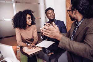 Three black individuals working together, one is explaining a topic using hand gestures while the other two smile in understanding.