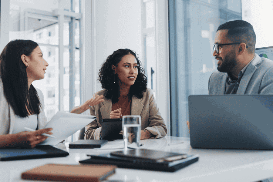 Three colleagues are sitting at a table in an office, discussing work. They look focused and engaged in their conversation. The image conveys a sense of professionalism and collaboration.