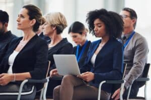 A group of business people seated in chairs, engaged in work-related activities. The setting appears to be indoors, with one featured woman using a laptop.