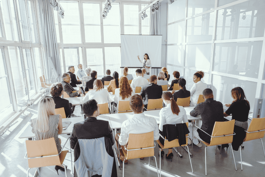 A diverse group of individuals seated in chairs around 3 tables, attentively facing a large screen in a meeting or presentation setting.