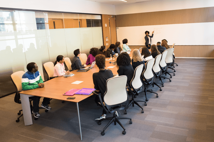 People sitting around a conference room table listening to woman at front of room pointing to whiteboard.