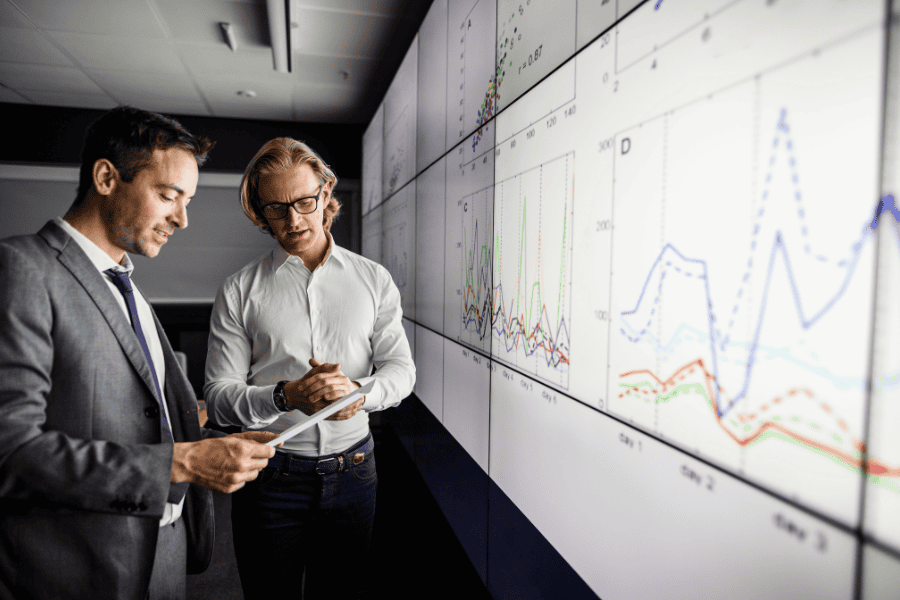 Two men in business suits analyzing graphs displayed on a large screen in a professional setting.
