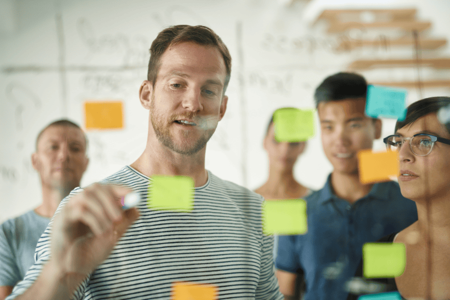 Man pointing at sticky notes on a window with coworkers standing behind him.