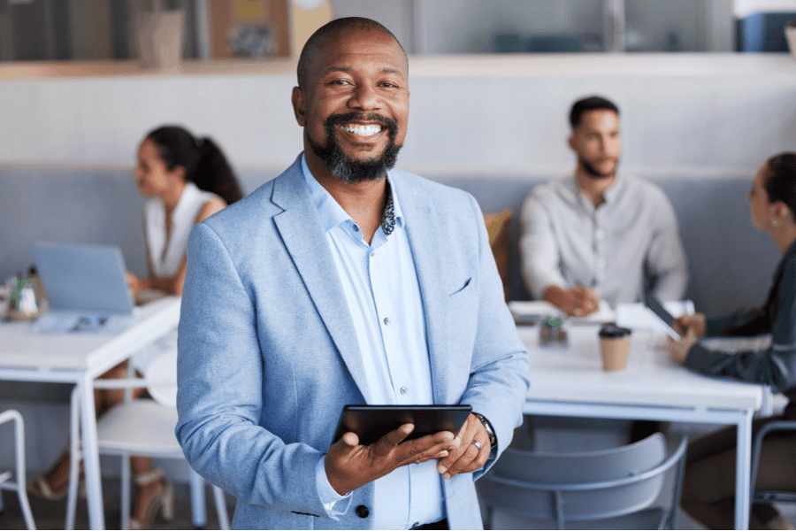 A man with a beard, dressed in blue, smiles at the camera while holding a tablet.