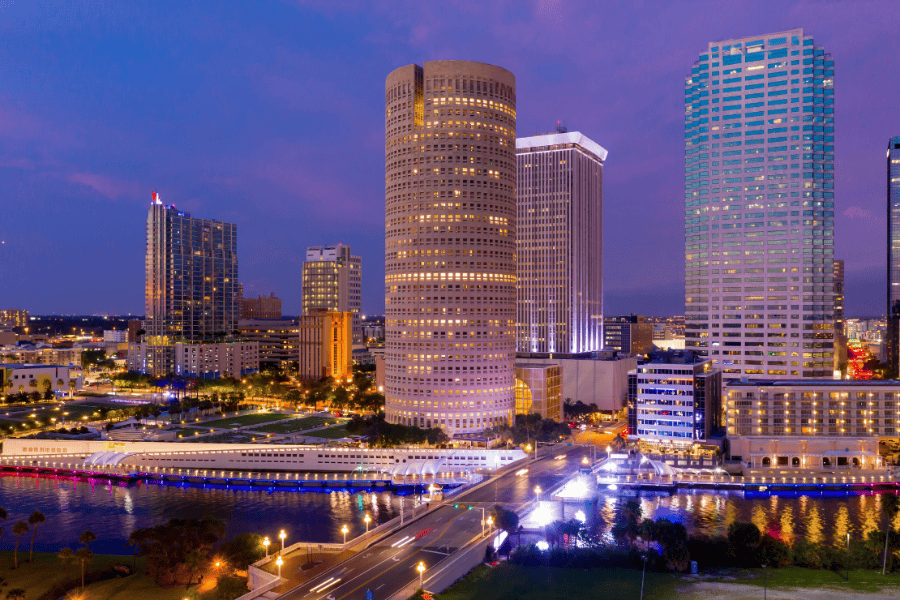 City skyline at dusk with a river and a bridge, reflecting warm colors in the water below.
