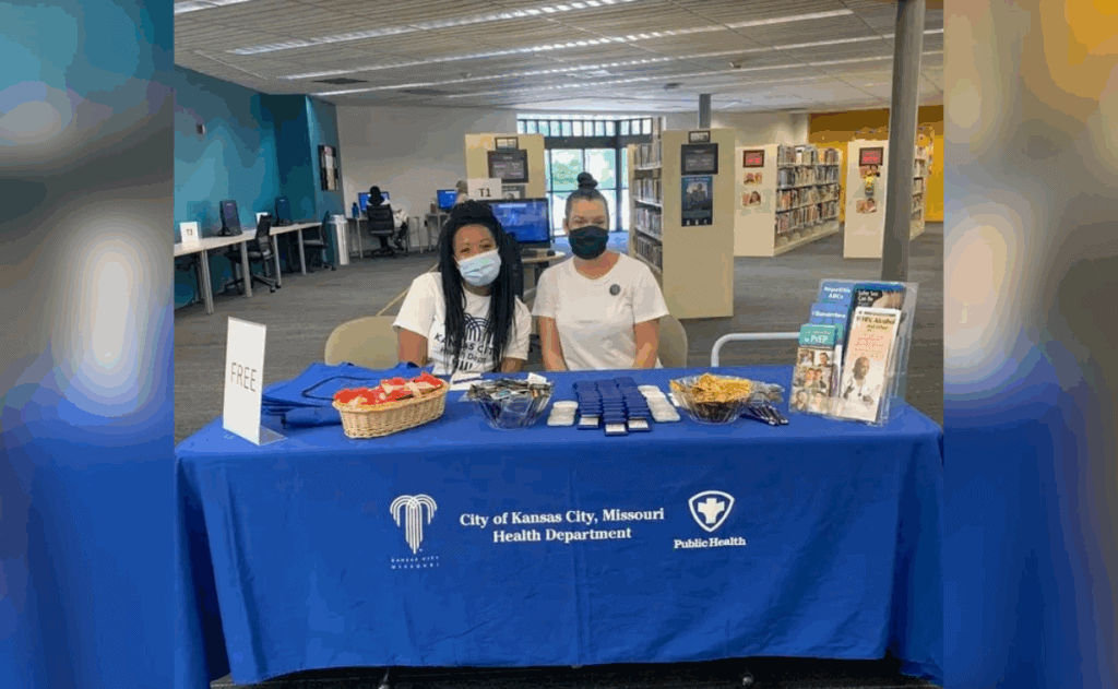Two workers that are wearing masks sit at a City of Kansas City, Missouri Health Department branded table