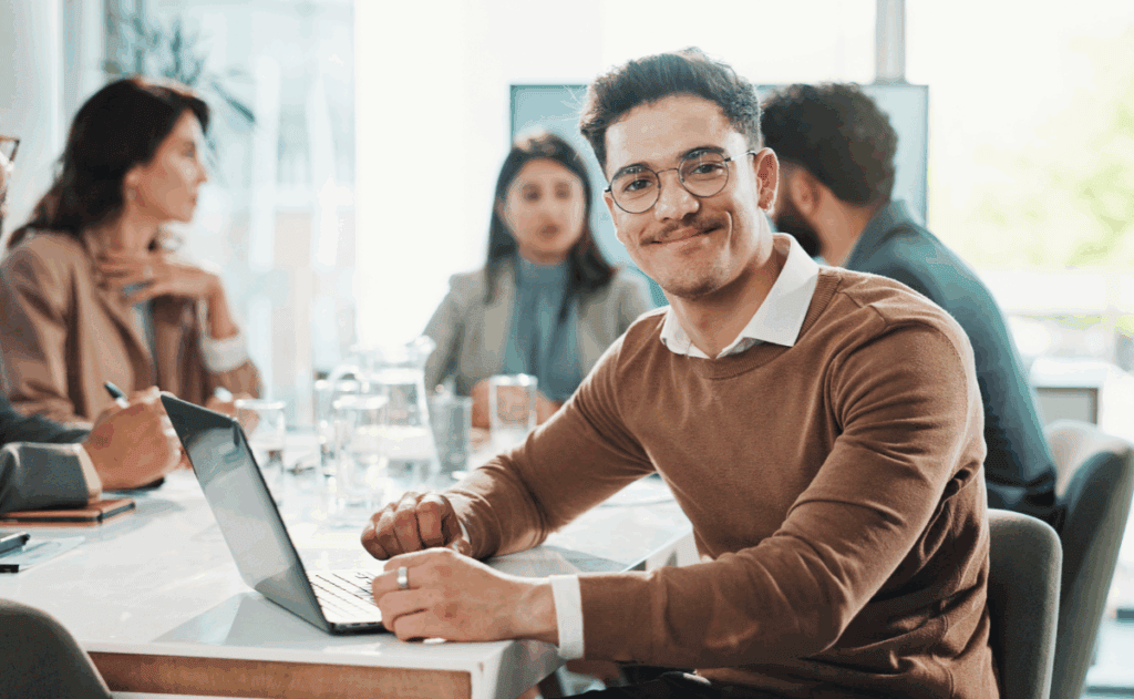 Man in glasses and a business professional outfit sitting at meeting table and smiling directly at the camera.