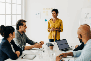 Woman in a yellow shirt standing at the head of a table during a meeting smiling with coworkers.