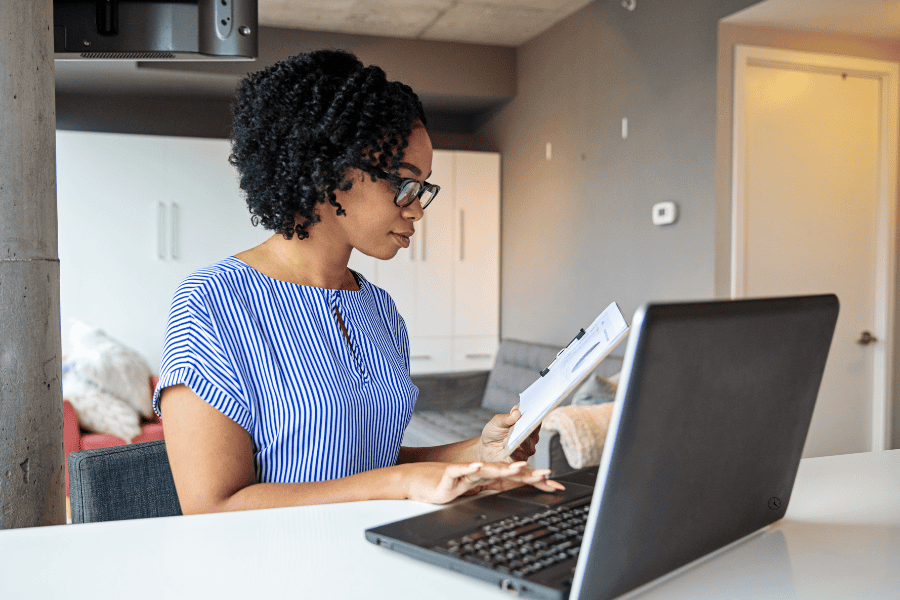 A person wearing glasses and a blue and white striped shirt works on a laptop while holding a document.