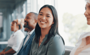 A group of people are sitting in a row, with a woman in the foreground smiling directly at the camera.