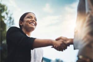 A woman smiles as she shakes hands with another person against a bright, cloudy sky.