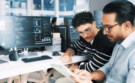 Two colleagues collaborating on a tablet in front of a computer screen displaying code.