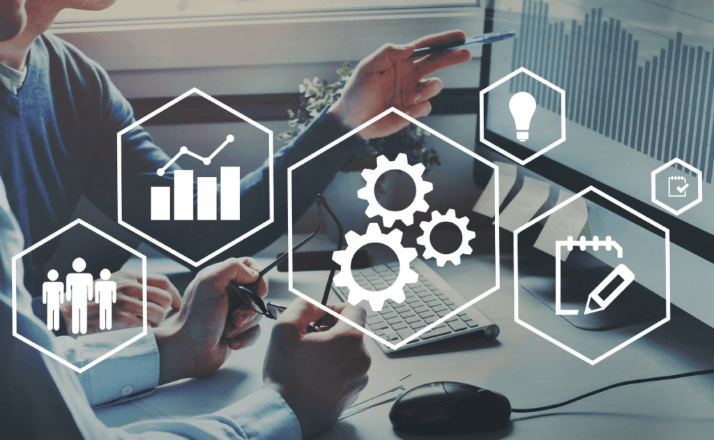 Two individuals are looking at a computer screen displaying a graph. Overlaid on the image are hexagonal icons representing teamwork, analytics, gears, ideas, and notes.