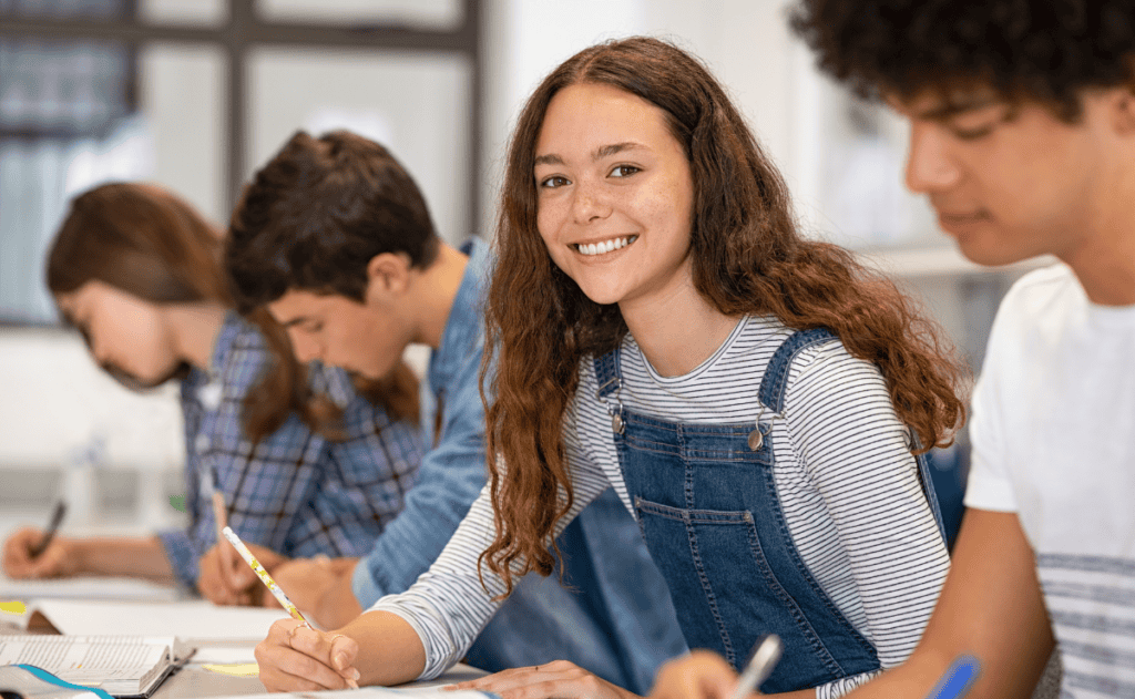 A student smiles at the camera while others focus on their work in a classroom setting.