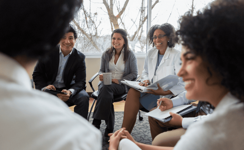 A group of professionals in a meeting are seated in a circle, engaged in discussion.