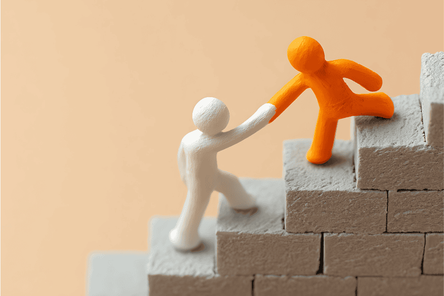 A white clay figure reaching up to help an orange clay figure climb stairs made of grey bricks.