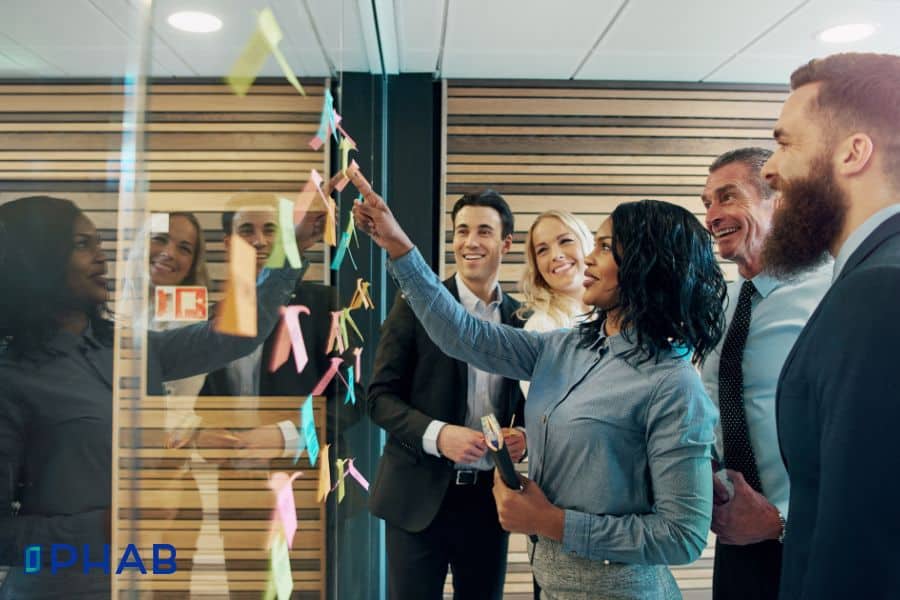A diverse group of colleagues collaborates around a glass wall covered in colorful sticky notes, with one person pointing and explaining ideas.
