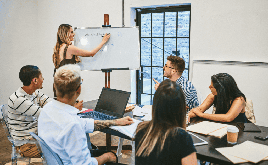 A group of people in a meeting room, with one person writing on a whiteboard and others looking on or engaged in discussion.