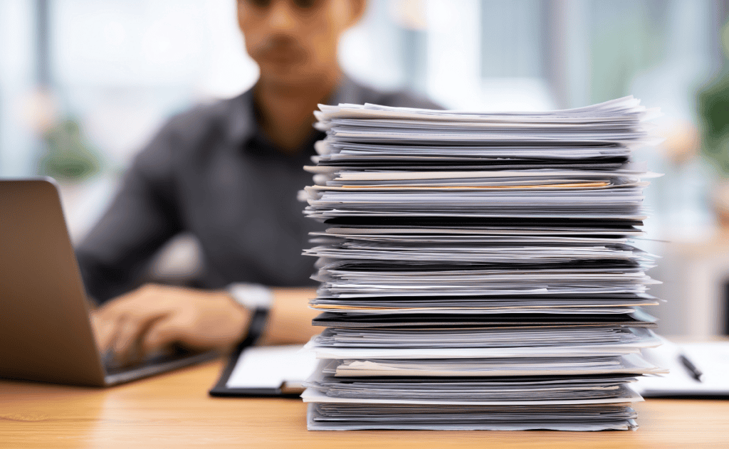 A stack of papers sits on a desk next to a person typing on a laptop.