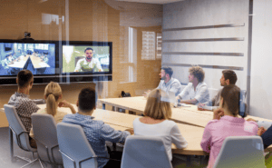 A group of people in a meeting room participate in a video conference, with two screens displaying participants and a live feed.