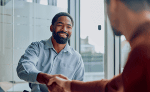 A man in a blue collared shirt smiles as he shakes hands with another person in an office setting.