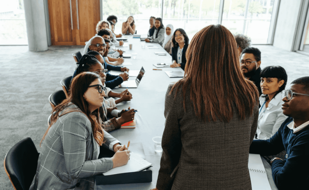 Team members sit around a conference table in a bright meeting room, taking notes and talking while one person in the foreground faces the group.
