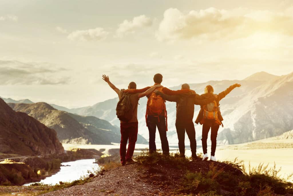 Five people stand on a mountain ridge by a winding river, arms outstretched in a group celebration as warm sunlight and distant snowcapped peaks fill the background.