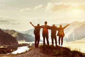 Five people stand on a mountain ridge by a winding river, arms outstretched in a group celebration as warm sunlight and distant snowcapped peaks fill the background.