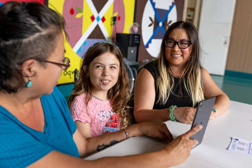 Three people sit at a table indoors, smiling during a conversation; an adult holds a tablet while a child and another adult look on, with colorful wall art in the background.