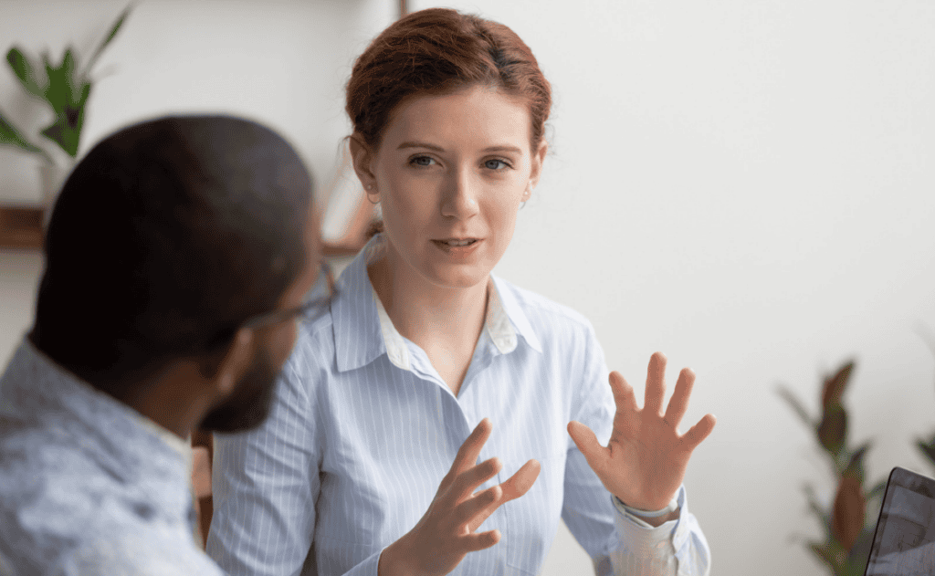 A woman in a light blue striped shirt speaks with a seated person across a table, gesturing with open hands during a discussion in a bright office.